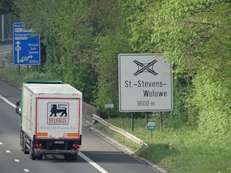 A truck with the logo and website of Delhaize is driving on a highway. The road is surrounded by lush green trees. A large road sign indicates the distance to St.-Stevens-Woluwe as 1600 meters. Another sign shows directions to cities like Brussels, Luik, Leuven, and Antwerp via the E40.