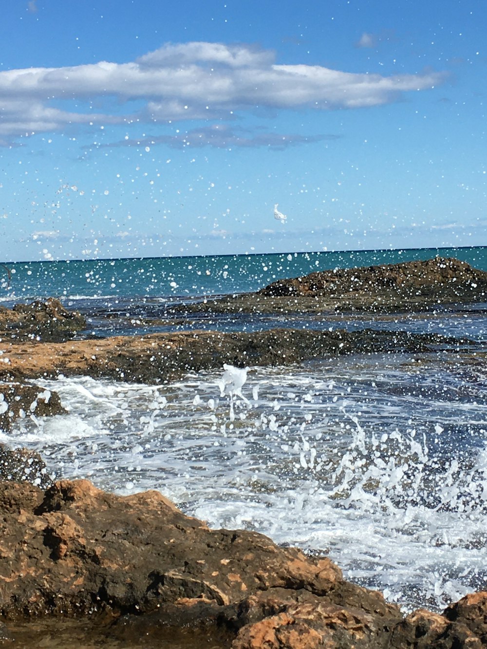 ocean waves crashing on brown rocky shore under blue and white cloudy sky during daytime