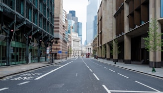 A wide, empty urban street lined with modern office buildings and a few young trees. The scene features a clear sky with some skyscrapers visible in the background. There is also a red double-decker bus in the distance, suggesting a city setting, possibly London.