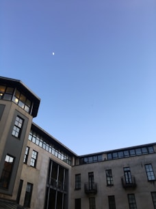 A partially visible moon in a clear blue sky above a multi-story building with modern design elements, including tall windows and small balconies. The structure has a combination of textures and neutral colors, creating a stark contrast with the sky.