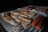 Grilled fish fillets and sausages are cooking over an open fire. The fish is seasoned, resting on a mesh grill with glowing coals underneath, indicating the food is being barbecued outdoors.