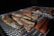 Grilled fish fillets and sausages are cooking over an open fire. The fish is seasoned, resting on a mesh grill with glowing coals underneath, indicating the food is being barbecued outdoors.