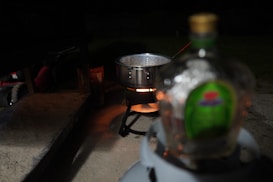 A cooking setup at night with a large pot on a portable stove emitting a warm glow. A blurred bottle with a green label is prominently in the foreground, slightly out of focus. The background is dimly lit, with hints of outdoor elements.