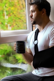 A man with a beard and short hair looks pensively out of a window while holding a mug with educational text. He is wearing a white t-shirt, grey pants, and has his right arm in a black sling. The background outside the window is lush with green foliage, suggesting a natural or garden setting.