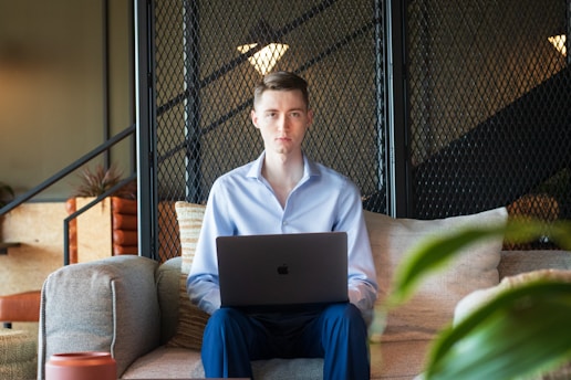 A person with short hair wearing a light blue shirt is sitting on a beige sofa with a laptop on their lap. The background features a black metal mesh partition and some indoor plants. The setting appears to be a modern indoor space with soft ambient lighting.