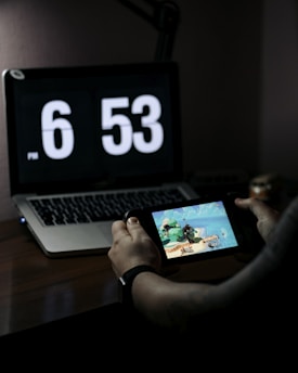 A person is holding a handheld gaming console while sitting at a desk. A laptop with a large digital clock display showing the time 6:53 PM is situated on the desk. The lighting is dim, creating a focused atmosphere.