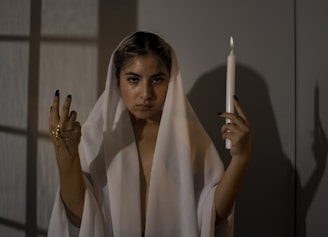 A person draped in a white cloth stands in a dimly lit room, holding a lit candle in one hand and a golden cross in the other. Shadows and soft lighting add to the dramatic and mysterious atmosphere.