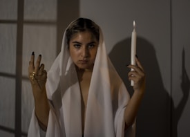 A person draped in a white cloth stands in a dimly lit room, holding a lit candle in one hand and a golden cross in the other. Shadows and soft lighting add to the dramatic and mysterious atmosphere.