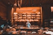 A cozy bakery with warm lighting, featuring shelves full of a variety of breads and pastries. A person in a baker's uniform is working at a counter, with glass display cases showing more baked goods. The walls and ceiling have a wooden design, adding a rustic charm to the space.