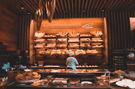 A cozy bakery with warm lighting, featuring shelves full of a variety of breads and pastries. A person in a baker's uniform is working at a counter, with glass display cases showing more baked goods. The walls and ceiling have a wooden design, adding a rustic charm to the space.