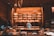 A cozy bakery with warm lighting, featuring shelves full of a variety of breads and pastries. A person in a baker's uniform is working at a counter, with glass display cases showing more baked goods. The walls and ceiling have a wooden design, adding a rustic charm to the space.