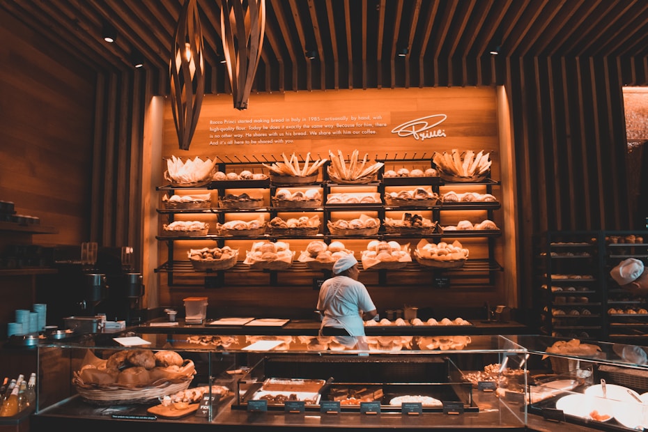 A cozy bakery with warm lighting, featuring shelves full of a variety of breads and pastries. A person in a baker's uniform is working at a counter, with glass display cases showing more baked goods. The walls and ceiling have a wooden design, adding a rustic charm to the space.