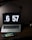 A dimly lit workspace featuring a laptop with a large digital clock display reading 6:57. In front of the laptop sits a handheld gaming console displaying a colorful game screen. The setup is on a dark wooden desk with a desk lamp providing subtle lighting.
