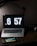 A dimly lit workspace featuring a laptop with a large digital clock display reading 6:57. In front of the laptop sits a handheld gaming console displaying a colorful game screen. The setup is on a dark wooden desk with a desk lamp providing subtle lighting.