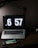 A dimly lit workspace featuring a laptop with a large digital clock display reading 6:57. In front of the laptop sits a handheld gaming console displaying a colorful game screen. The setup is on a dark wooden desk with a desk lamp providing subtle lighting.