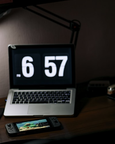 A dimly lit workspace featuring a laptop with a large digital clock display reading 6:57. In front of the laptop sits a handheld gaming console displaying a colorful game screen. The setup is on a dark wooden desk with a desk lamp providing subtle lighting.
