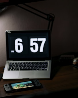 A dimly lit workspace featuring a laptop with a large digital clock display reading 6:57. In front of the laptop sits a handheld gaming console displaying a colorful game screen. The setup is on a dark wooden desk with a desk lamp providing subtle lighting.