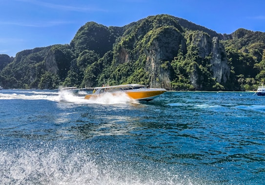 A vibrant yellow speedboat moves quickly through the water, creating a splash and wake behind it. The surrounding area features lush, green, rocky cliffs under a clear blue sky, indicating a tropical or coastal setting.