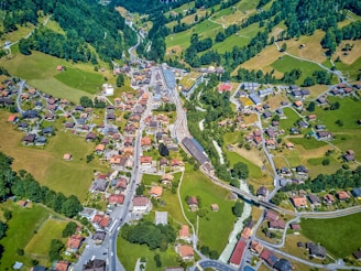 An aerial view of a small village nestled in a hilly landscape. The scene features numerous houses with red and brown rooftops scattered along winding roads. Lush greenery and patches of farmland surround the village, with forests blanketing the hills in the background. A railway cuts through the center of the image, adding an element of connectivity.