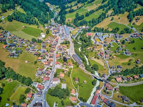 An aerial view of a small village nestled in a hilly landscape. The scene features numerous houses with red and brown rooftops scattered along winding roads. Lush greenery and patches of farmland surround the village, with forests blanketing the hills in the background. A railway cuts through the center of the image, adding an element of connectivity.