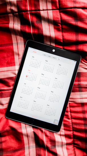 A digital tablet displaying a calendar for the year 2020. The tablet is placed on a red plaid fabric surface, creating a contrast between technology and textiles.