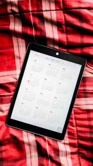 A digital tablet displaying a calendar for the year 2020. The tablet is placed on a red plaid fabric surface, creating a contrast between technology and textiles.