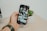 A hand holding a smartphone displaying an Instagram profile with multiple images, beside a small potted succulent plant on a light wooden desk.