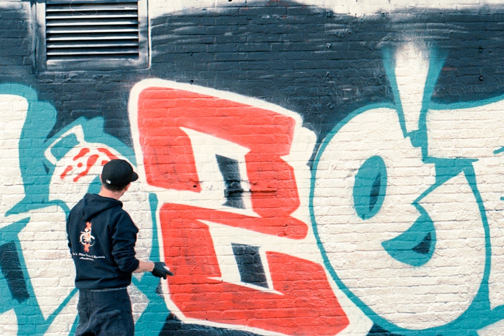 man in gray jacket standing beside wall with graffiti