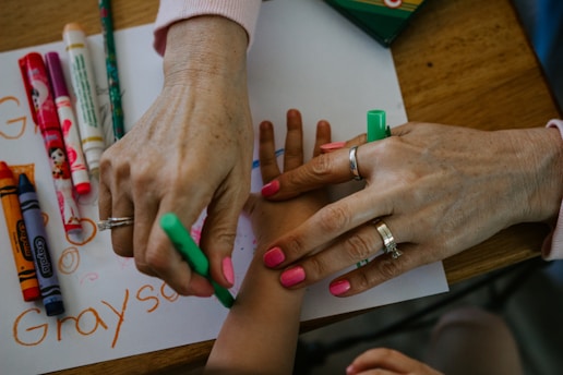 Two hands are visible, one adult and one child, with the adult hand drawing on the child's arm using a green marker. Various colorful markers and crayons are spread across the wooden table, and a sheet of paper with writing and drawings is partially visible.