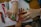 Two hands are visible, one adult and one child, with the adult hand drawing on the child's arm using a green marker. Various colorful markers and crayons are spread across the wooden table, and a sheet of paper with writing and drawings is partially visible.