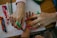 Two hands are visible, one adult and one child, with the adult hand drawing on the child's arm using a green marker. Various colorful markers and crayons are spread across the wooden table, and a sheet of paper with writing and drawings is partially visible.