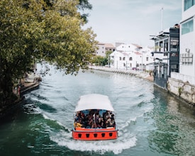 A red and white boat is traveling along a narrow river or canal, surrounded by urban buildings. On the left, large green trees overhang the water, while on the right, modern white and gray buildings line the waterfront. Several people are seated in the boat, some taking photographs.