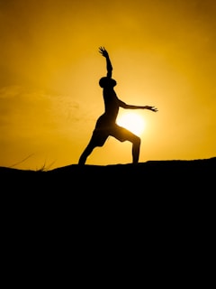 A silhouette of a person striking a dynamic yoga pose against the backdrop of a glowing sun. The sky is a warm, golden hue, creating a dramatic contrast with the dark figure.