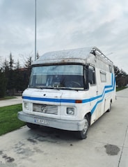 A vintage camper van with a white and blue color scheme is parked on a concrete path. The van has a worn and rustic look, with some visible rust and wear marks on the exterior. In the background, tall trees and a cloudy sky can be seen.