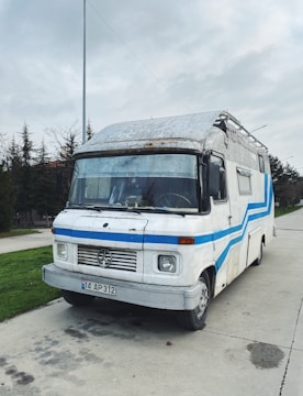 A vintage camper van with a white and blue color scheme is parked on a concrete path. The van has a worn and rustic look, with some visible rust and wear marks on the exterior. In the background, tall trees and a cloudy sky can be seen.