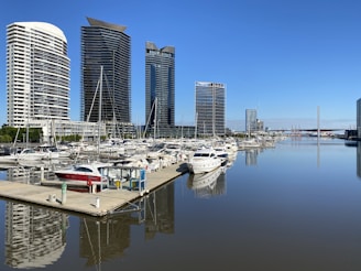 Several modern high-rise buildings line the edge of a marina, with numerous boats and yachts docked on still water. The clear blue sky and calm waters provide a serene backdrop for the urban landscape.