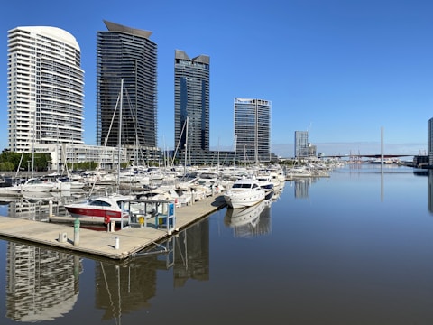 Several modern high-rise buildings line the edge of a marina, with numerous boats and yachts docked on still water. The clear blue sky and calm waters provide a serene backdrop for the urban landscape.