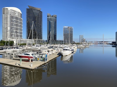 Several modern high-rise buildings line the edge of a marina, with numerous boats and yachts docked on still water. The clear blue sky and calm waters provide a serene backdrop for the urban landscape.
