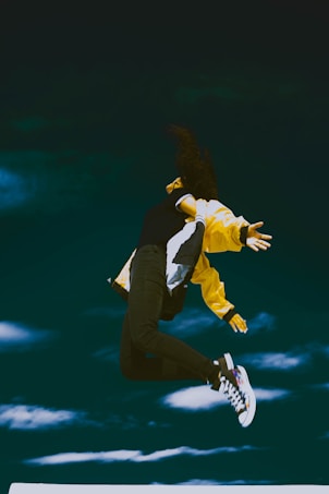 A person is captured mid-air against a deep teal sky, exhibiting a vibrant and dynamic jump. They are wearing a black top, dark pants, and a bright yellow jacket, with stylish, colorful sneakers. The person