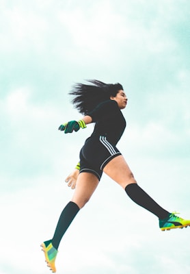 A person appears to be in mid-air, dressed in athletic attire with long socks, cleats, and neon-accented gloves. The background is a bright, cloudy sky, conveying a sense of motion and athleticism.