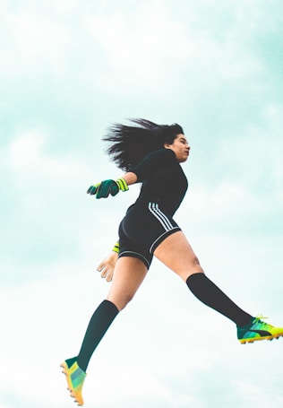 A person appears to be in mid-air, dressed in athletic attire with long socks, cleats, and neon-accented gloves. The background is a bright, cloudy sky, conveying a sense of motion and athleticism.