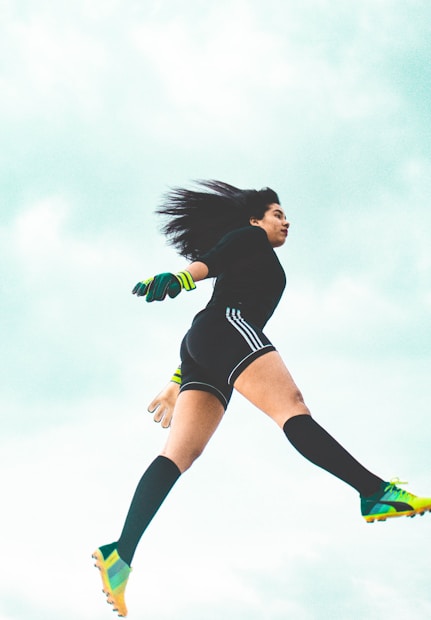 A person appears to be in mid-air, dressed in athletic attire with long socks, cleats, and neon-accented gloves. The background is a bright, cloudy sky, conveying a sense of motion and athleticism.