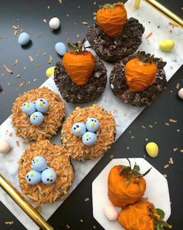 A variety of themed desserts arranged on a white surface, including donuts topped with orange chocolate-coated strawberries and donuts resembling bird nests with blue candy eggs. Crushed cookies and toasted coconut flakes are used for decoration. Small pastel-colored eggs scatter around the scene.