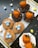 A variety of themed desserts arranged on a white surface, including donuts topped with orange chocolate-coated strawberries and donuts resembling bird nests with blue candy eggs. Crushed cookies and toasted coconut flakes are used for decoration. Small pastel-colored eggs scatter around the scene.