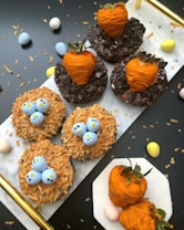 A variety of themed desserts arranged on a white surface, including donuts topped with orange chocolate-coated strawberries and donuts resembling bird nests with blue candy eggs. Crushed cookies and toasted coconut flakes are used for decoration. Small pastel-colored eggs scatter around the scene.