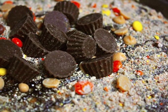 A close-up view of chocolatey treats and candies surrounded by a powdery mix. Dark brown chocolate cups are prominently scattered at the center, accompanied by colorful candy-coated chocolates and light brown round candies.