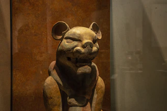 A sculpture of an animal with features resembling a bear or feline, crafted from a material that gives a rustic appearance. The figure has rounded ears, closed eyes, and a serene expression standing against a brown textured background.