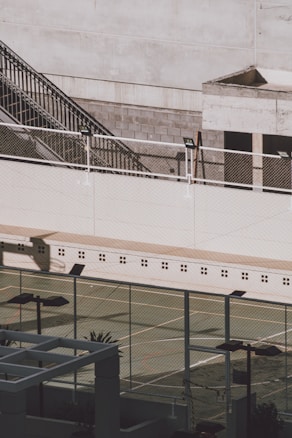 An outdoor sports court surrounded by a high, sturdy fence. There are sunlight and shadows cast onto the court's surface. A staircase and some industrial-looking structures are visible in the background.