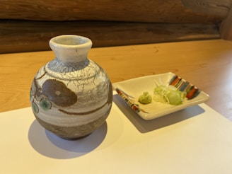 A ceramic jug with artistic patterns sits on a light wooden surface next to a square dish containing small portions of green wasabi and shredded white radish. The background includes a wooden log wall, adding to the rustic ambiance.