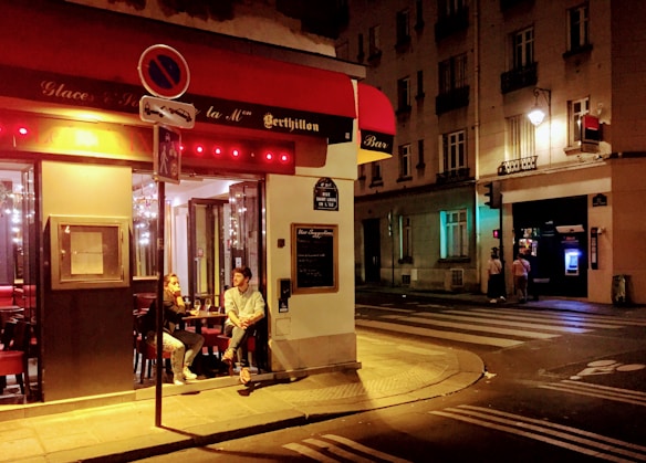 A cozy evening scene at a street corner caf&eacute; with two people seated at a small table outside. Warm lights illuminate the interior of the caf&eacute;, casting a welcoming glow on the surrounding sidewalk. Nearby, a streetlamp lights up the corner of a quiet urban setting, with additional pedestrians visible near an ATM across the street.
